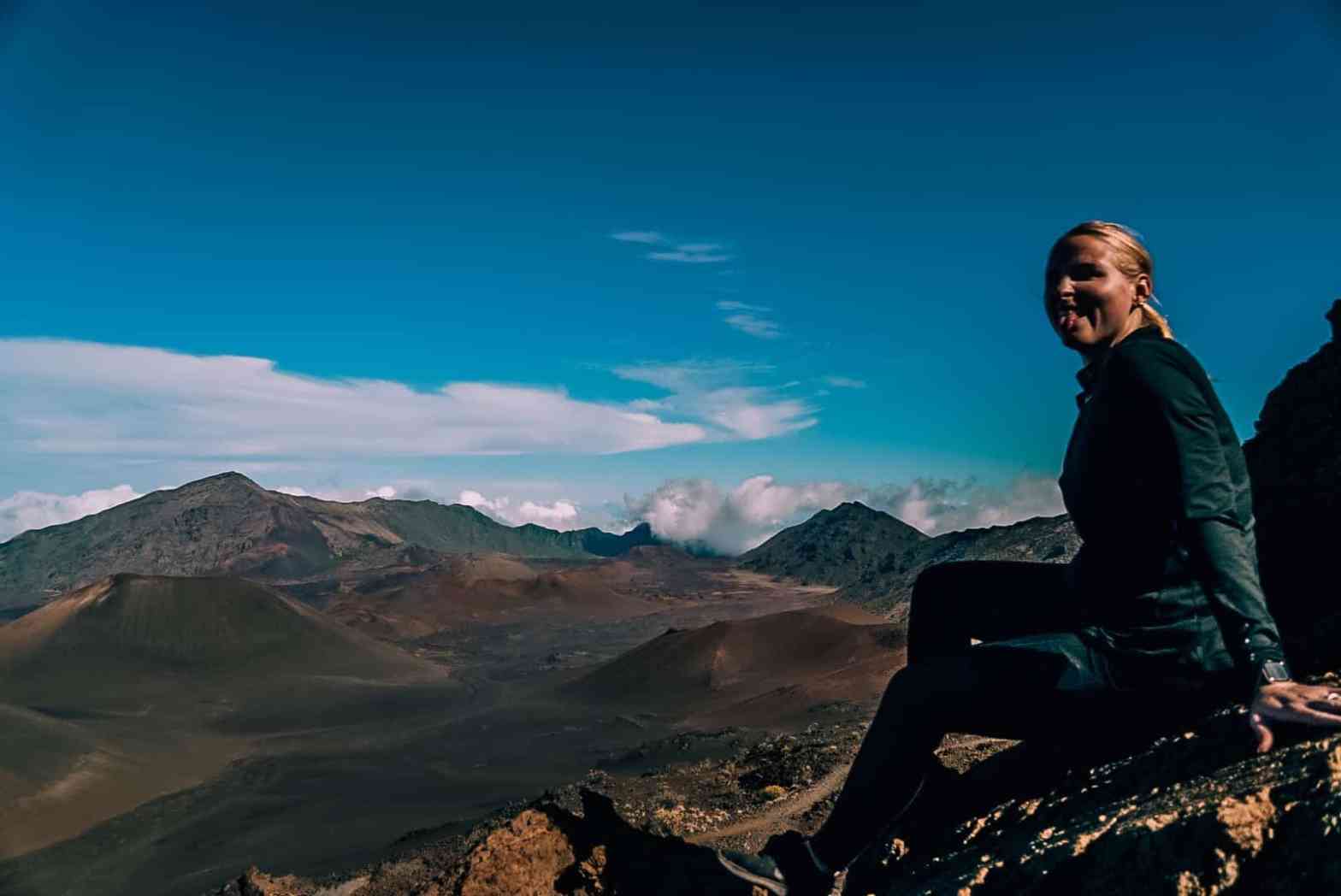 Woman sitting on a rock smiling with her tongue out in front of the Haleakala crater