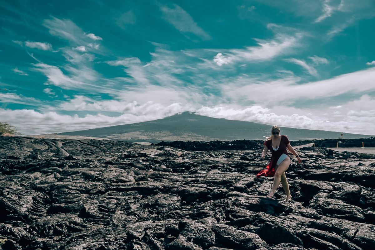 Woman in swimsuit and coverup standing on old lava rock bed with Mauna Loa in the background