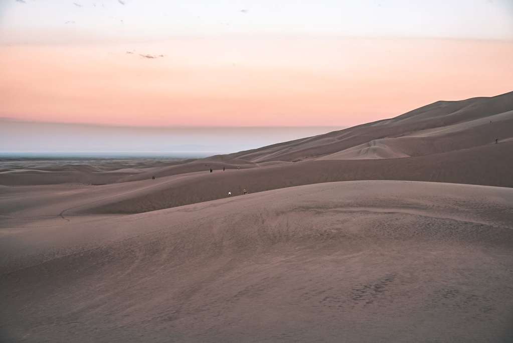 View of people on the Great Sand Dunes during sunrise with the sky pastel purple, pink, and yellow