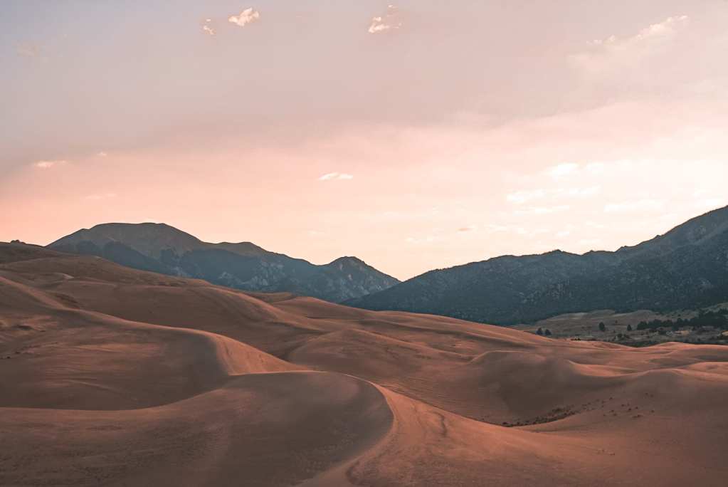 View of the Great Sand Dunes facing the mountains during sunrise with the sky pastel purple, pink, and yellow