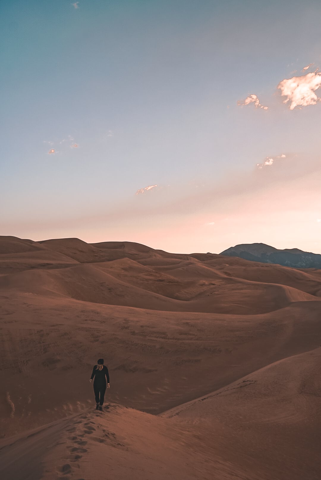 View of woman walking away onto a dune within the Great Sand Dunes during sunrise with the sky pastel pink and blue sky