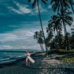 Woman walking in white swimsuit down a black sand beach with palm trees in the background and blue skies and water