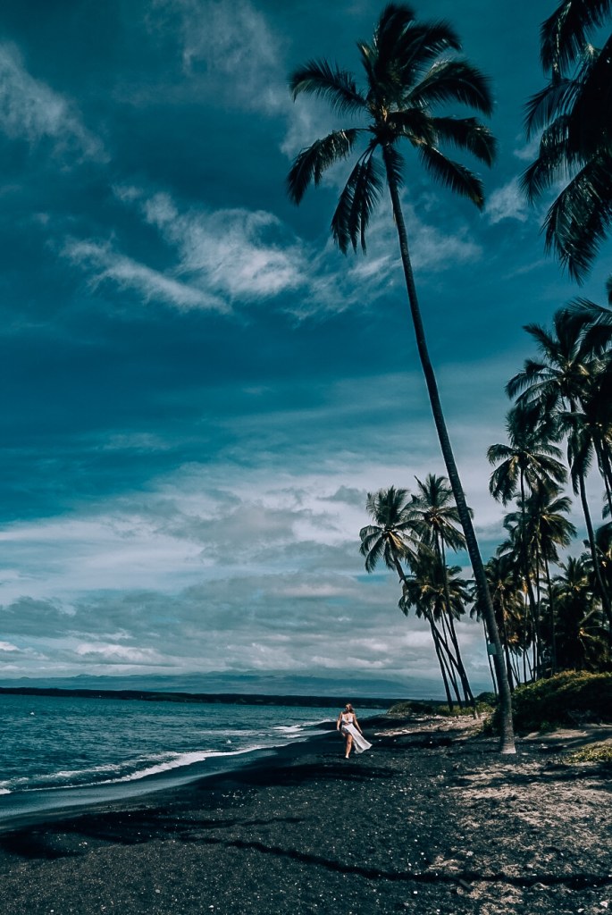 Woman walking in white swimsuit down a black sand beach with palm trees in the background and blue skies and water