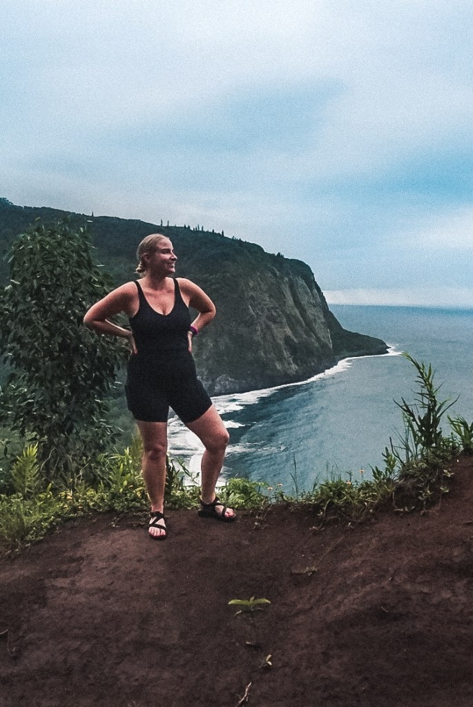 Woman in exercise tank top, shorts, and sandals standing in front of Waipio Valley from high up