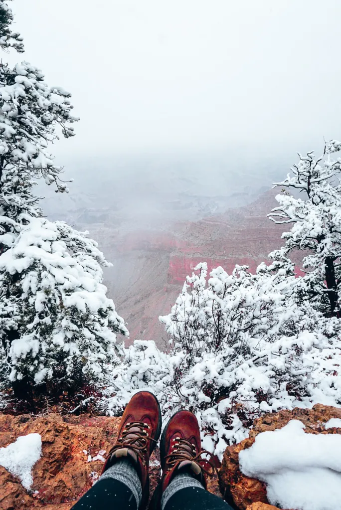 Woman's hiking boots and socks sitting on the edge of the Grand Canyon in front of the snow covered trees