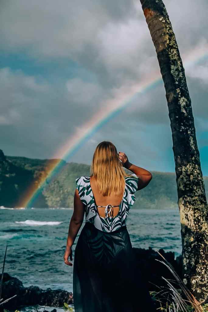 Woman standing in a swimsuit and skirt wrap next to a palm tree, looking out to the ocean to see a rainbow