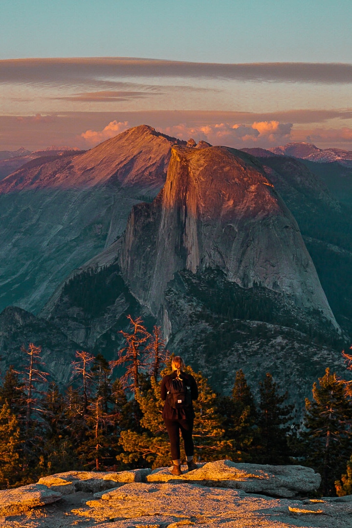 Woman standing atop Sentinel Dome looking out at Half Dome over sunset