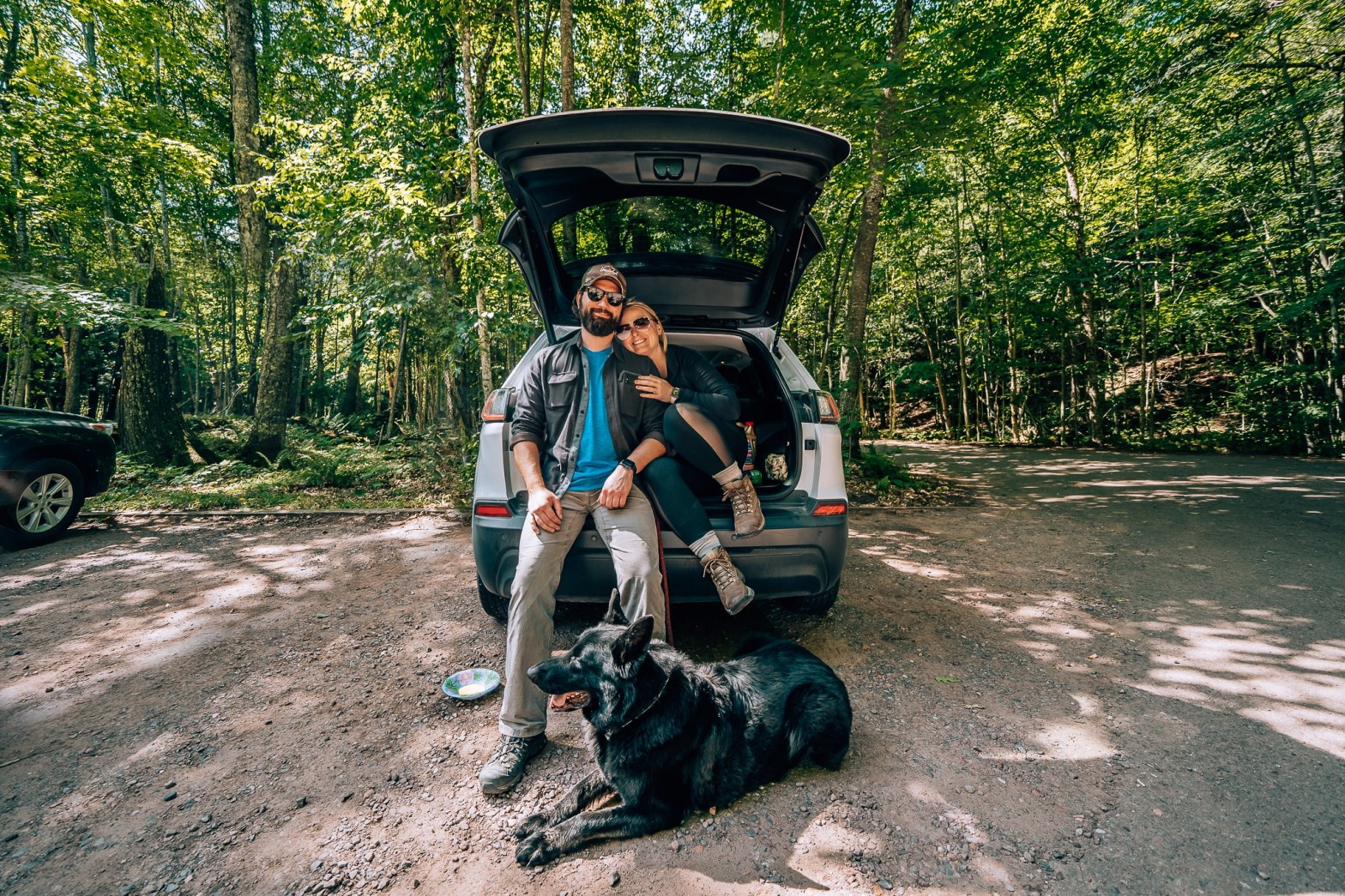 Man and woman sat in the back of a Jeep Cherokee with their black German Shepherd laying down in front during a road trip