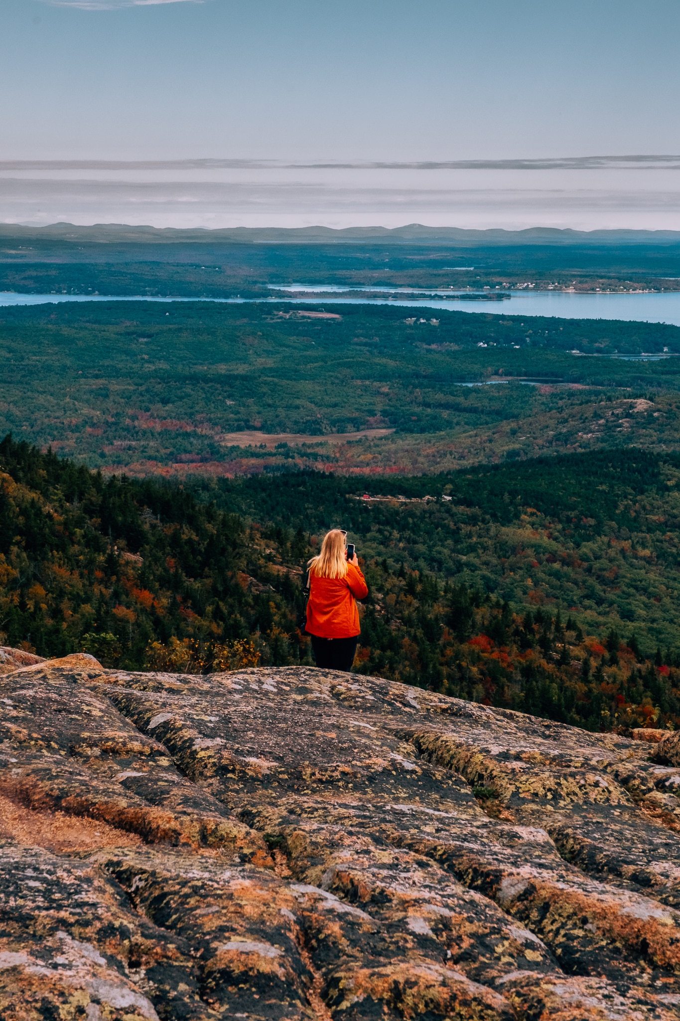 Woman standing atop a mountain looking out to the view with the coastline and many red and yellow trees for autumn