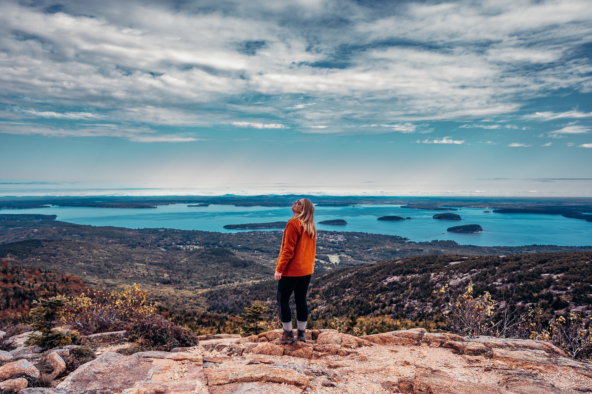 Woman standing atop a viewpoint with the coastline and Bar Harbor in the background