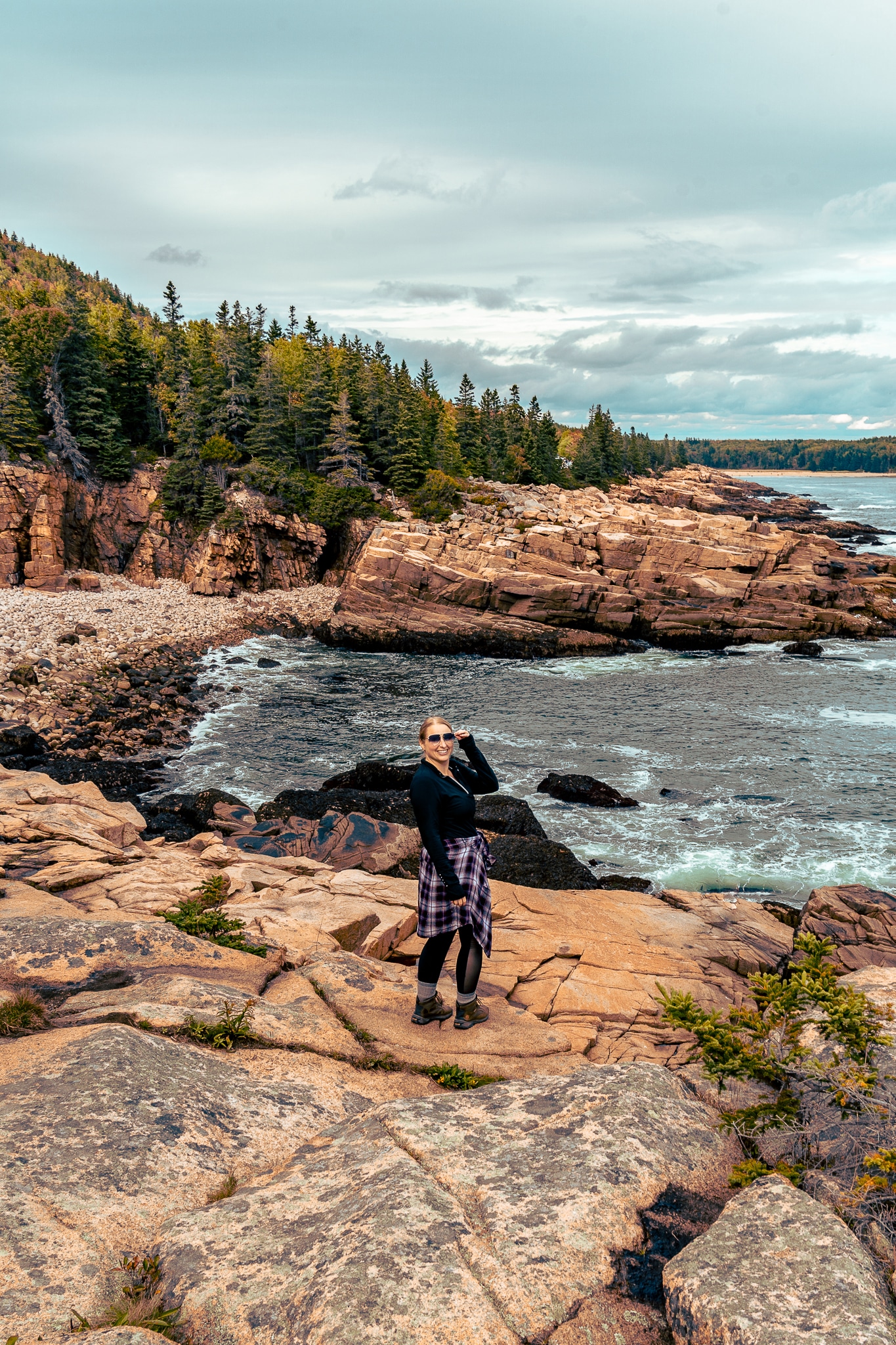 Woman standing at Schooner Head Overlook at Acadia National Park, with the coastline jagged behind her