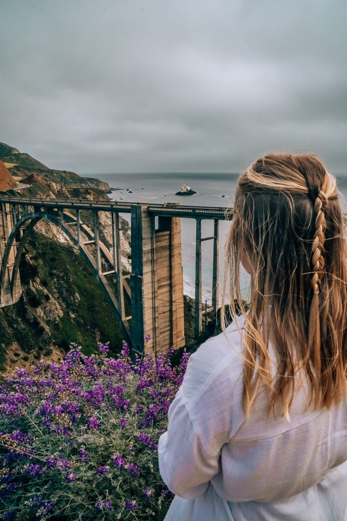 Woman standing in front of a purple flowered-bush looking out to Bixby Bridge in Big Sur along the California coastal highway