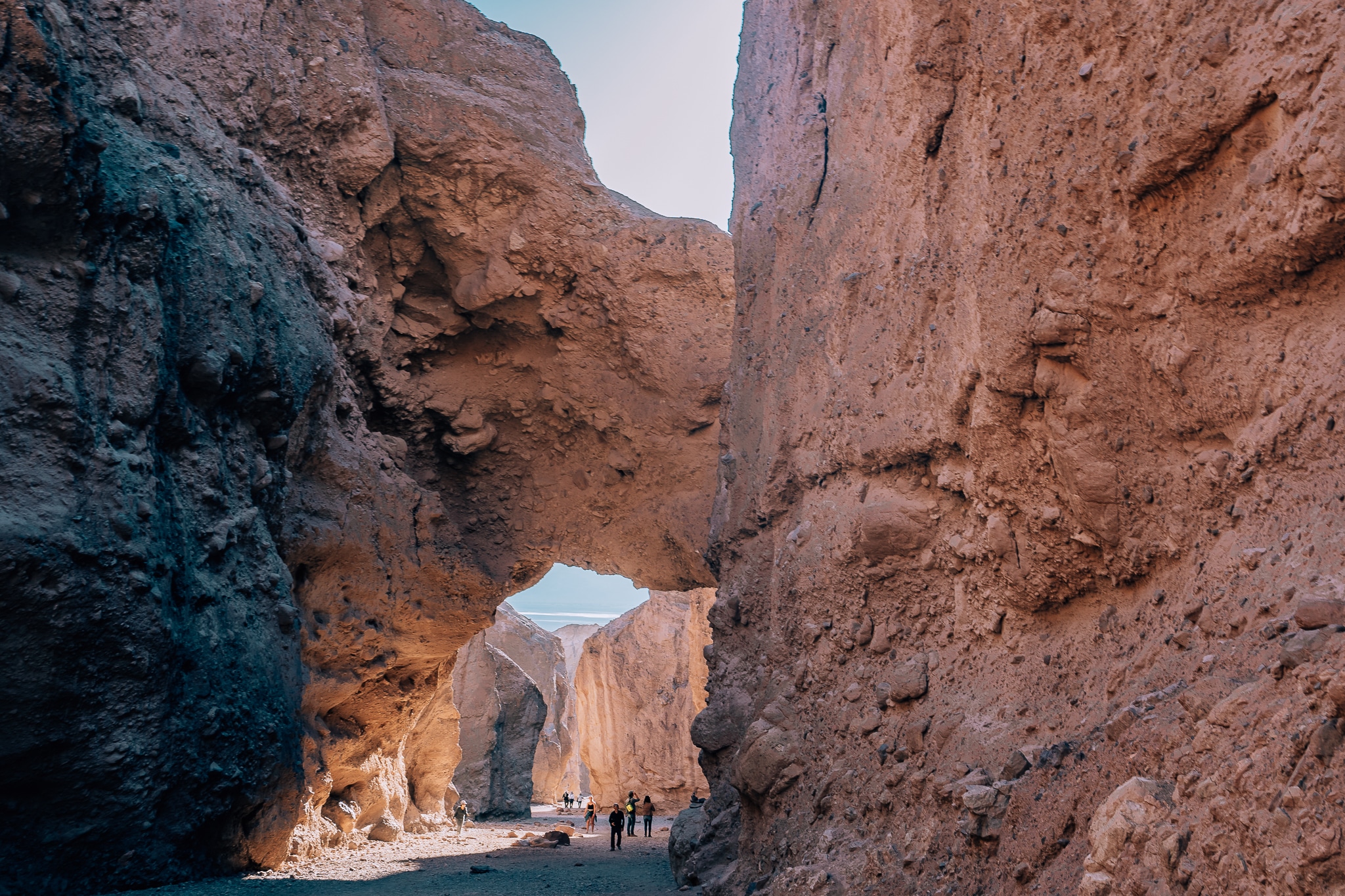 A large natural arch connecting two sides of a large orange canyon with people walking underneath at Death Valley