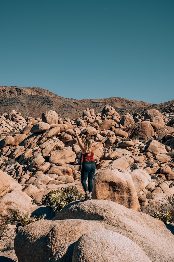 Woman with her arms up while standing on a Joshua Tree rock while in a field of rocks