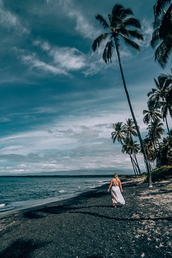 Woman walking along a black sand beach in a white swimsuit and coverup under the palm trees on the Big Island of Hawaii