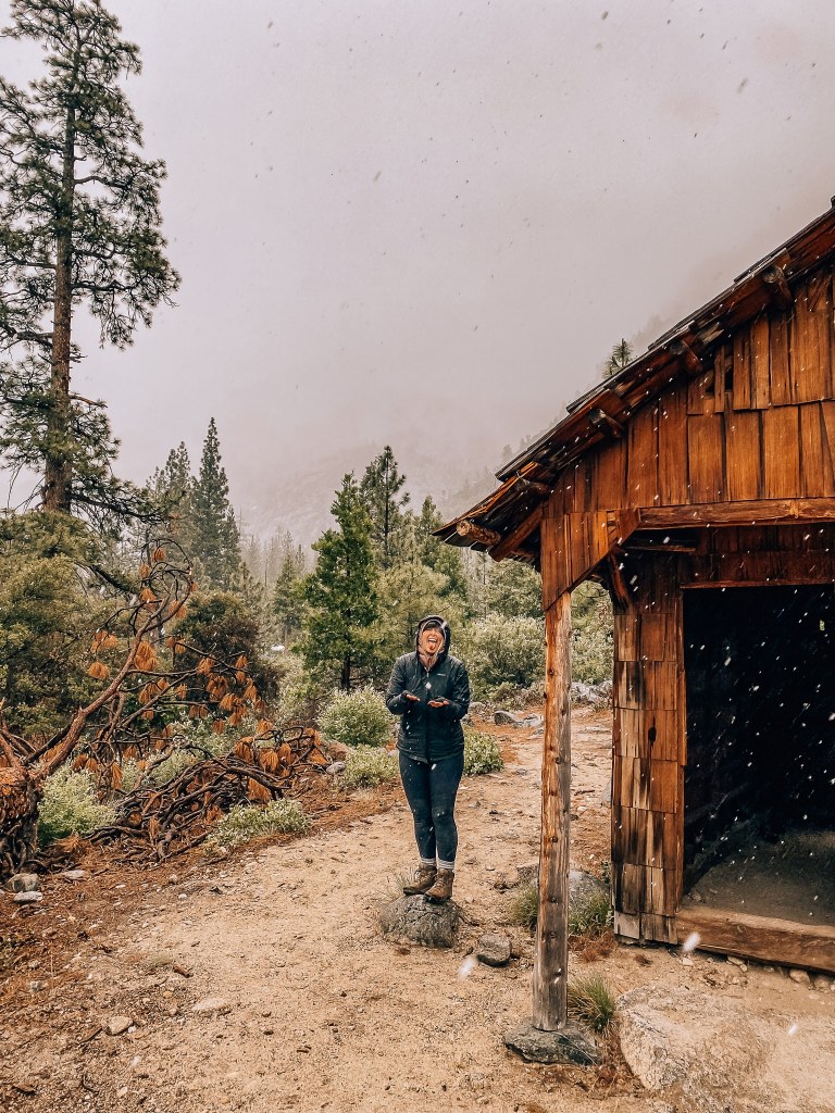 Woman standing next to a cabin with her tongue out while it snows