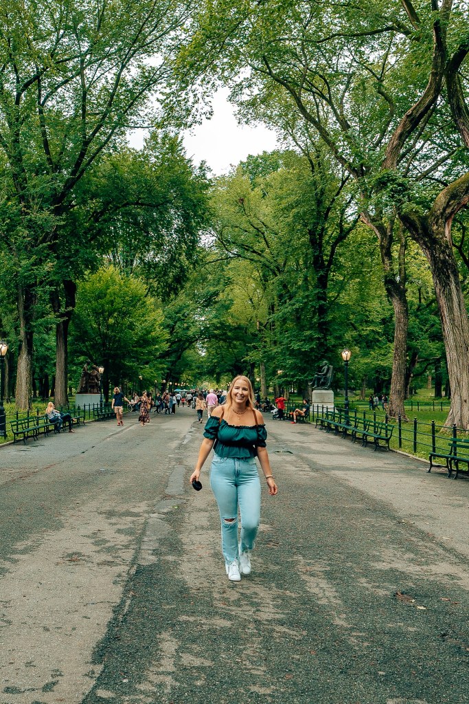 Smiling blonde woman in green shirt and jeans walking toward you in The Mall in Central Park with the tall, spiraling green trees behind her