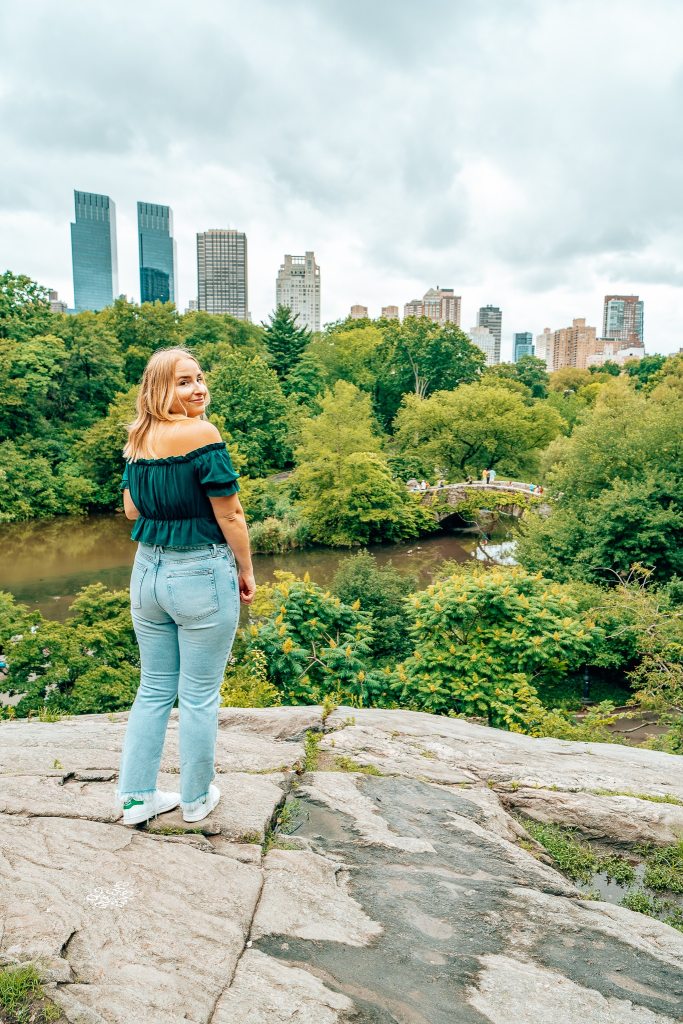 Woman standing on Umpire Rock in Central Park looking out toward the pond, with a bridge going over it, and the New York City skyline in the background