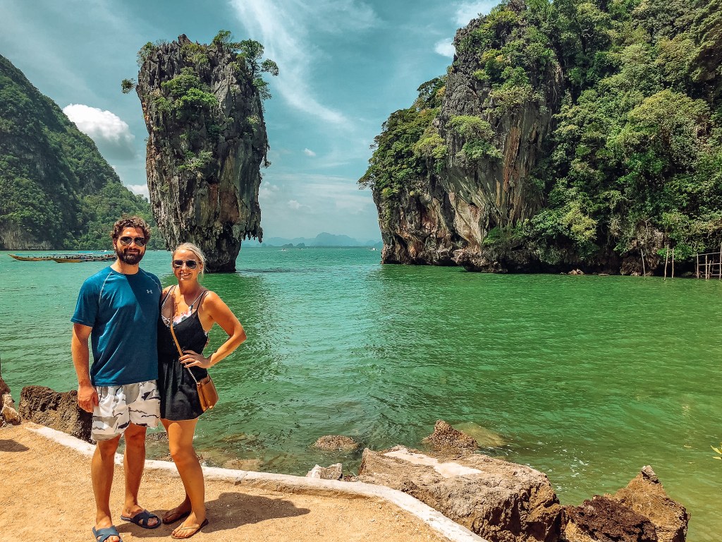Smiling man and woman standing in front of James Bond Island, a very small but tall island, sticking out of the water in Thailand