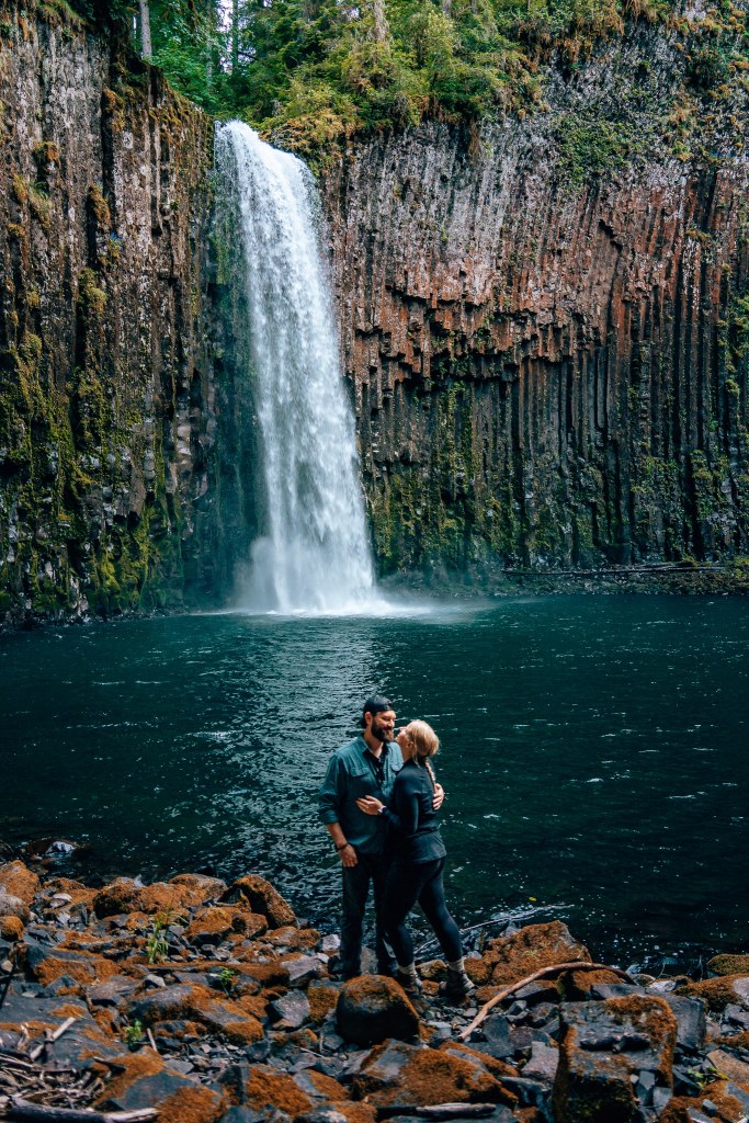 Man and woman embracing, looking into each others' eyes smiling in front of a waterfall