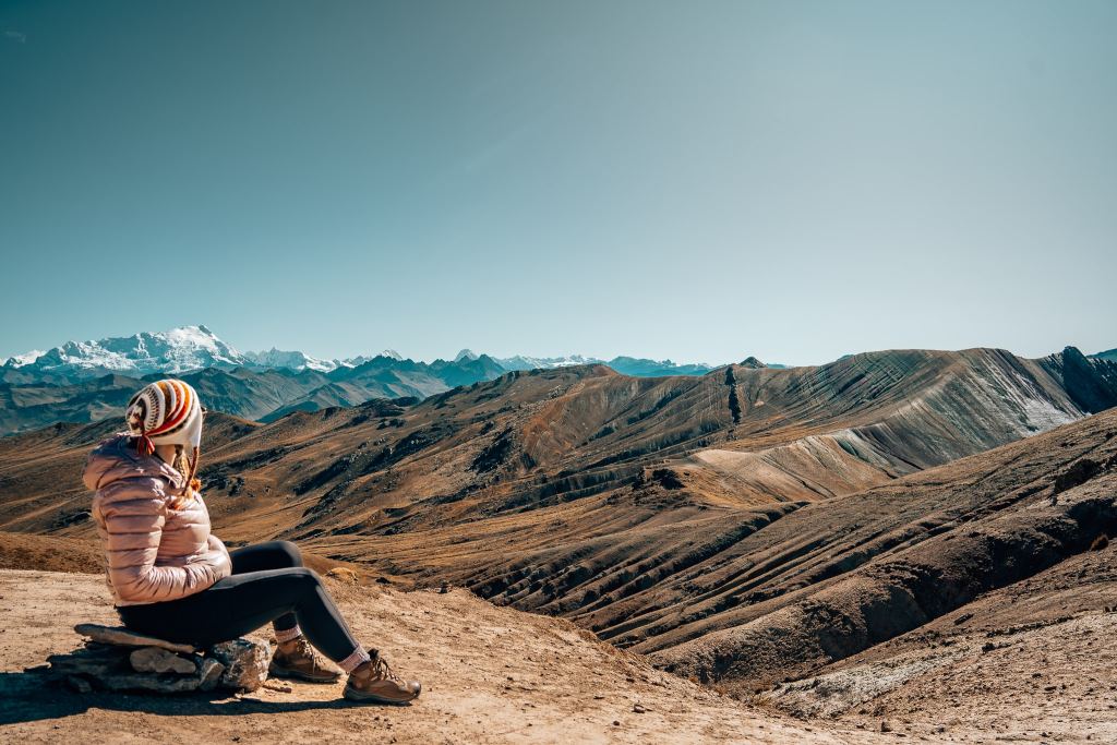 Woman sitting on a rock looking out to snow-capped mountains and also rainbow mountains