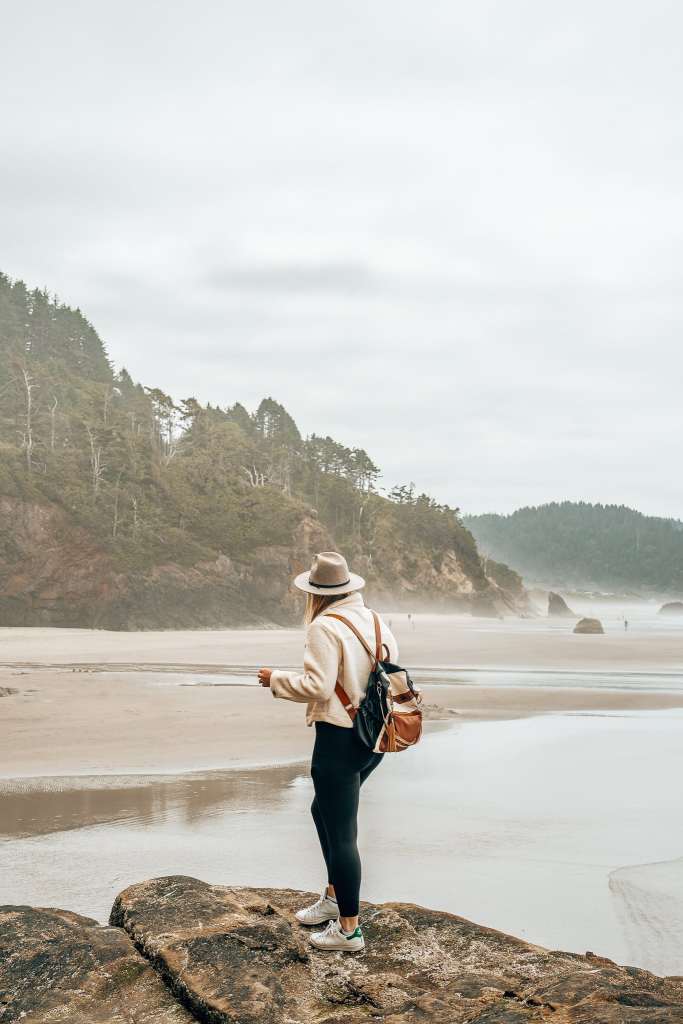Woman in a hat, jacket, and backpack standing on a rock looking out to a hazy Oregon coast
