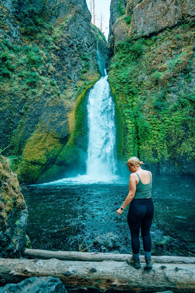 Woman standing on a fallen log in front of Wahclella Falls in Columbia River Gorge