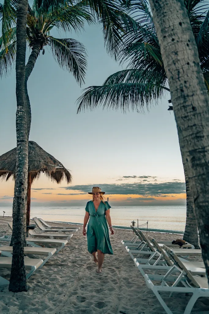 Woman wearing a hat and dress walking through daybeds on the beach with the sunrise in the background