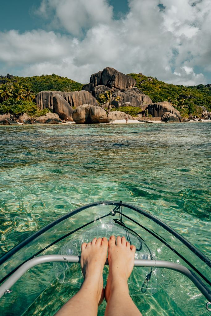 Woman sitting in a clear kayak with the sea below her and large granite boulders of La Digue in front