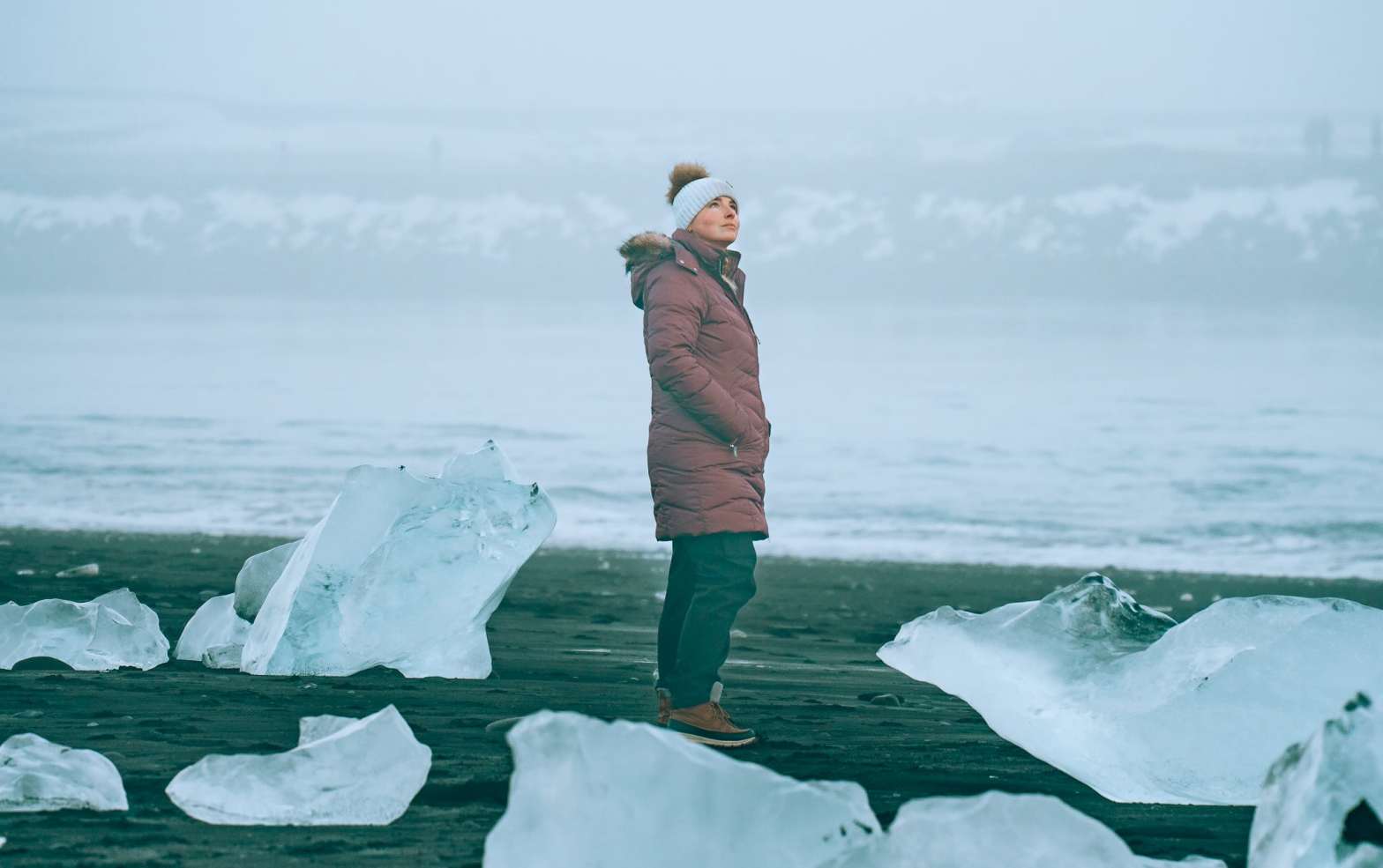 Woman in a coat and hat standing on Diamond Beach in Iceland, with the giant glacier chunks surrounding her on the beach
