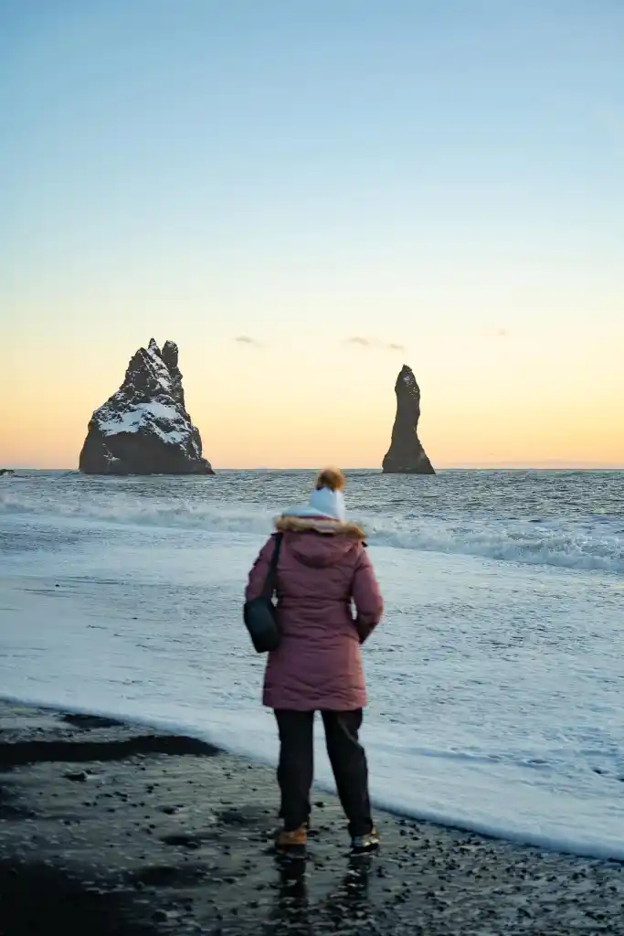 Woman standing on Reynisfjara black sand beach with the sun setting in the background