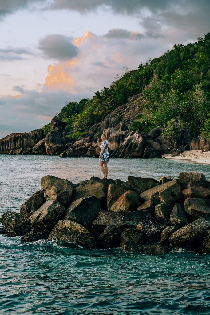 Woman standing on rocks jutting out into the water in the Seychelles with the sunset behind her