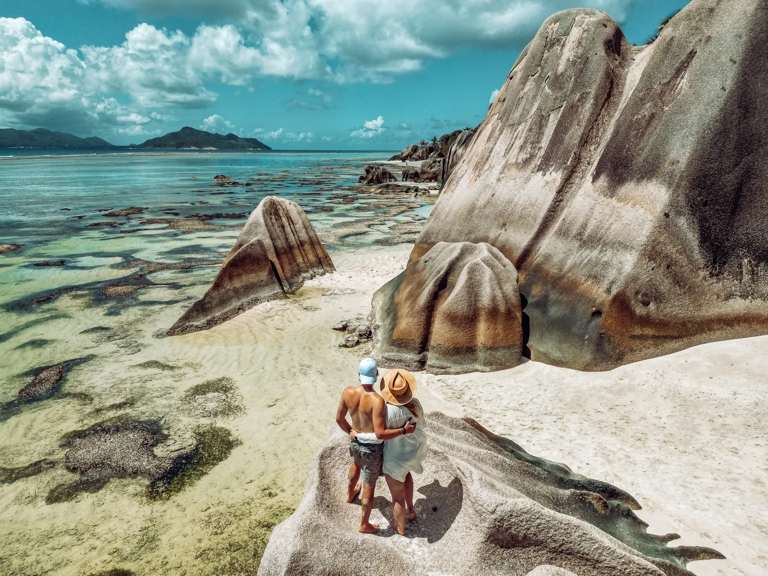 Man and woman standing on a rock looking out to the iconic beach of Anse Source d'Argent on La Digue in the Seychelles, covered in large granite boulders with the sea to the left