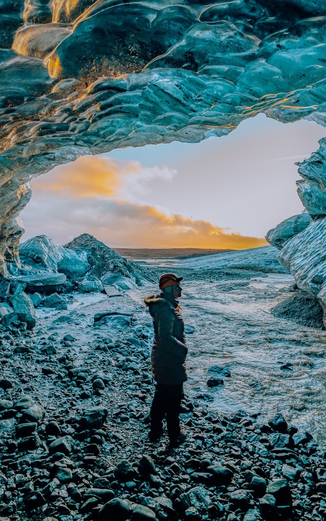 Woman standing in an ice cave with the sun setting in the entrance to the cave in the background