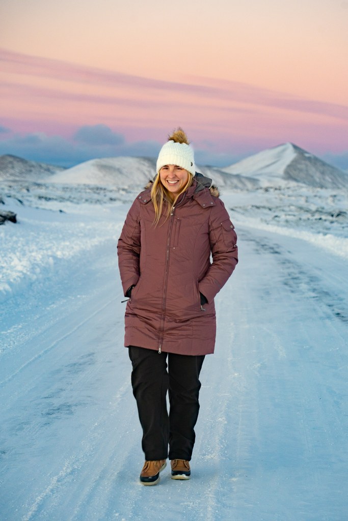Woman in a coat, hat, and other winter gear walking with snowy mountains and a sunrise sky in the background