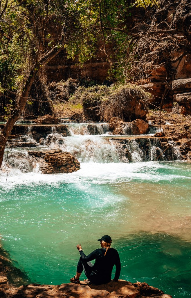 Woman sitting on the banks of a river with waterfalls cascading down in front of her at Havasupai