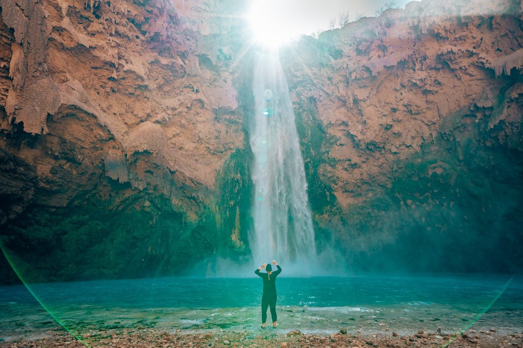 Woman standing at the base of Mooney Falls with the sun peaking out over top