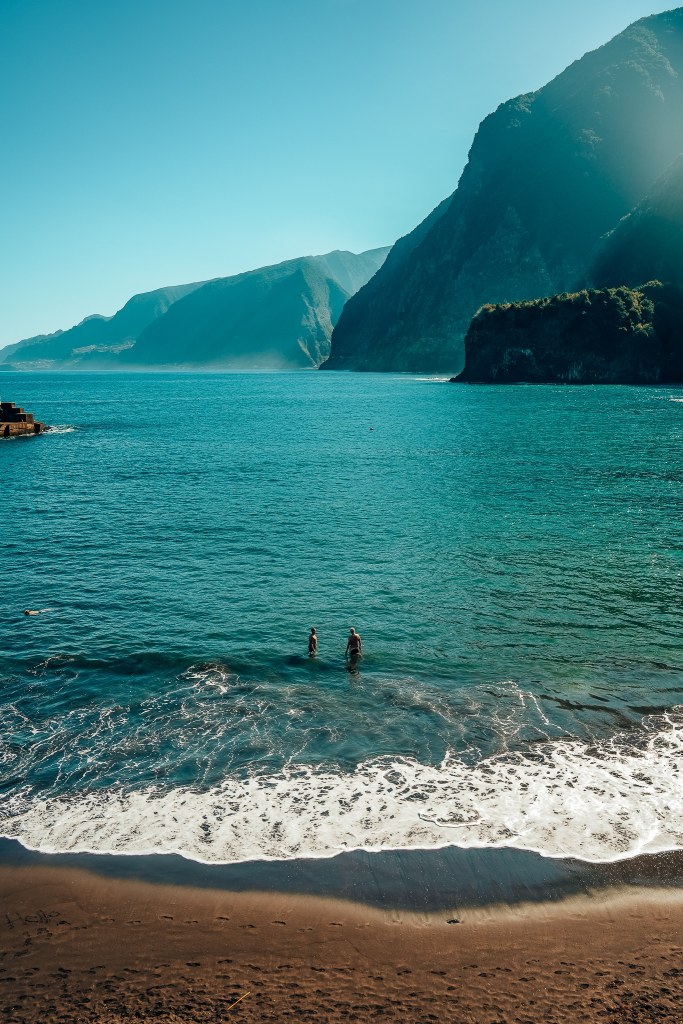 Two people standing in the water on a black sand beach in Madeira with cliffs in the background