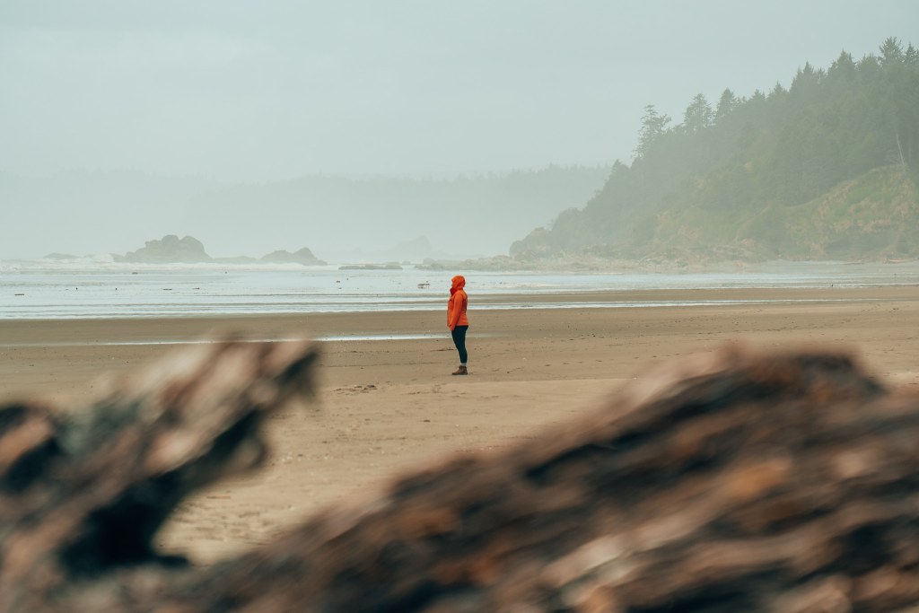 Woman standing on Kalaloch Beach at Olympic National Park with a fallen tree in the foreground, the woman in a bright raincoat standing far away, and the shoreline in the background