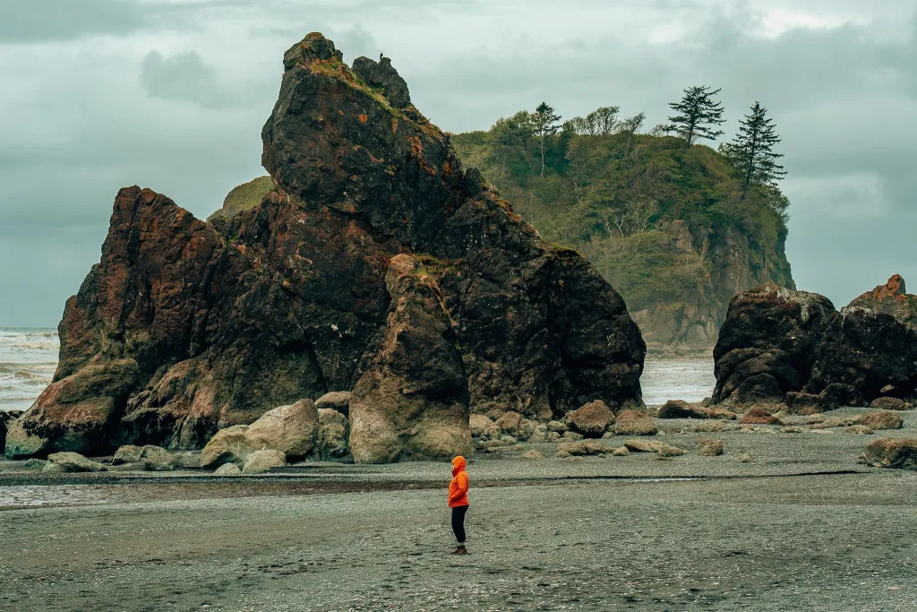 Woman in an orange raincoat standing on the beach in front of giant sea stacks at Ruby Beach in Olympic National Park