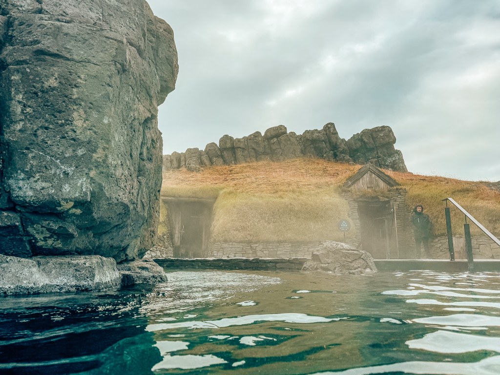 View from within the pool at Sky Lagoon looking up to a grass-roofed house and a security guard standing in front of it