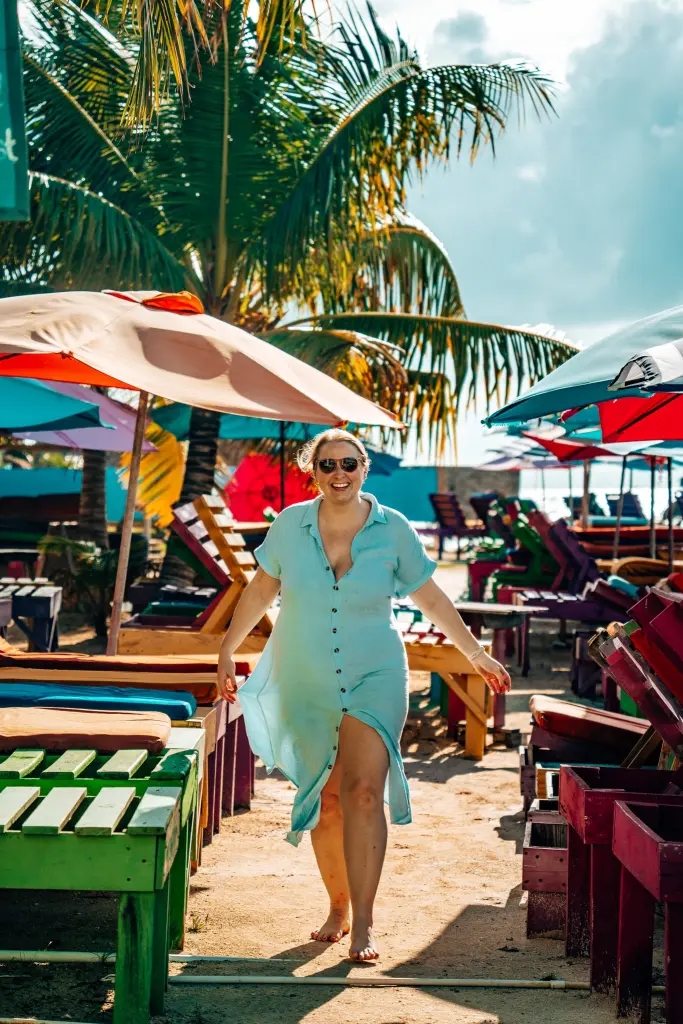 Woman in a blue swim cover up standing between beach lounge chairs and in front of an umbrella and palm trees at Secret Beach in Ambergris Caye, Belize