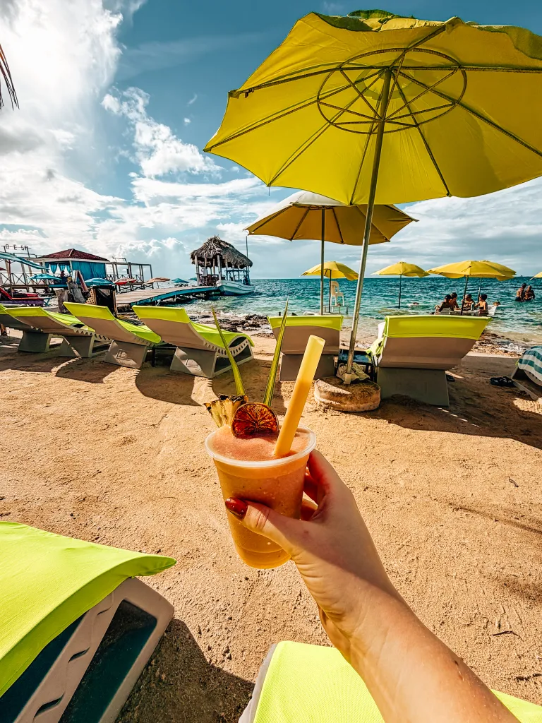 Hand holding a frozen strawberry cocktail with beach umbrellas and lounge chairs in the bakground