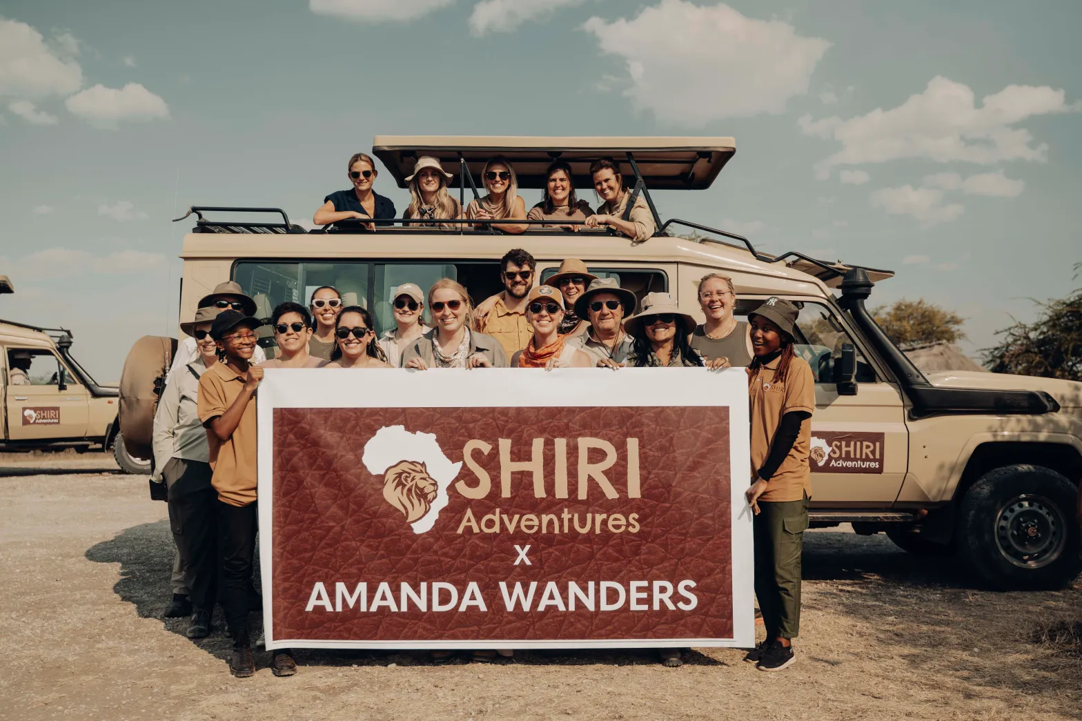 Group of 20 people in and around a safari jeep holding a sign that says Shiri Adventures x Amanda Wanders